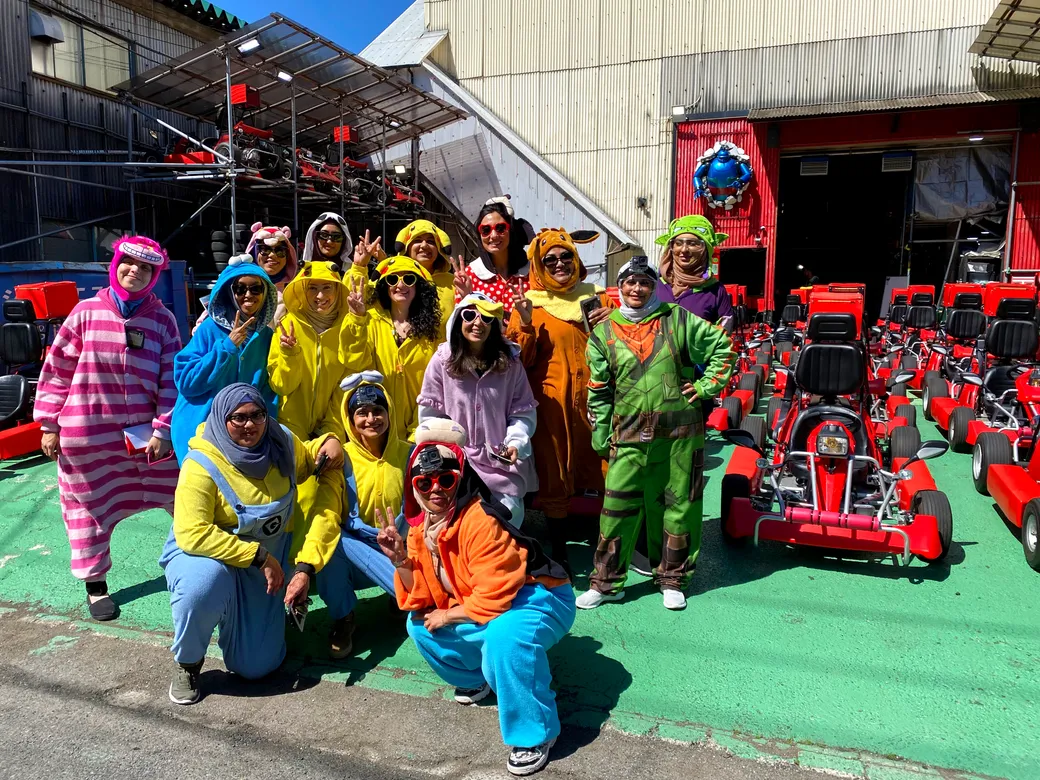 Group of people in bright costumes posing for a photo on a go-kart track, with red go-karts lined up behind them on a sunny day.