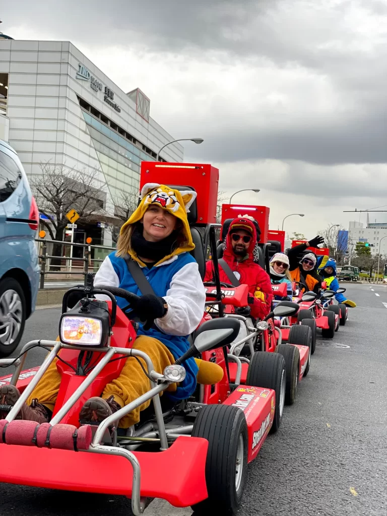 People in red pedal go-karts riding in a street parade, front rider wearing a tiger hoodie and blue overalls, smiling at the camera.