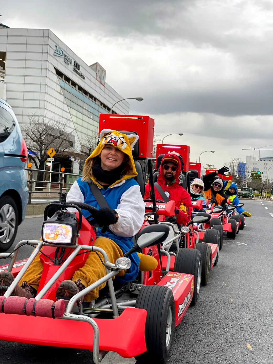 People in red pedal go-karts riding in a street parade, front rider wearing a tiger hoodie and blue overalls, smiling at the camera.