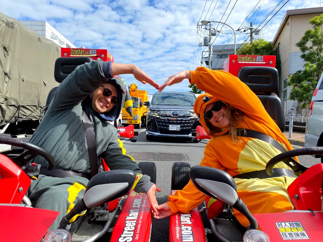 Two people in colorful racing outfits in go-karts high-five while smiling at the camera on a city street event.