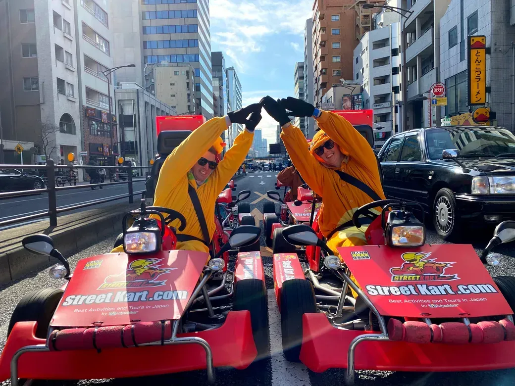 Two people in bright yellow outfits on red go-karts form a heart shape with their arms in a busy city street.
